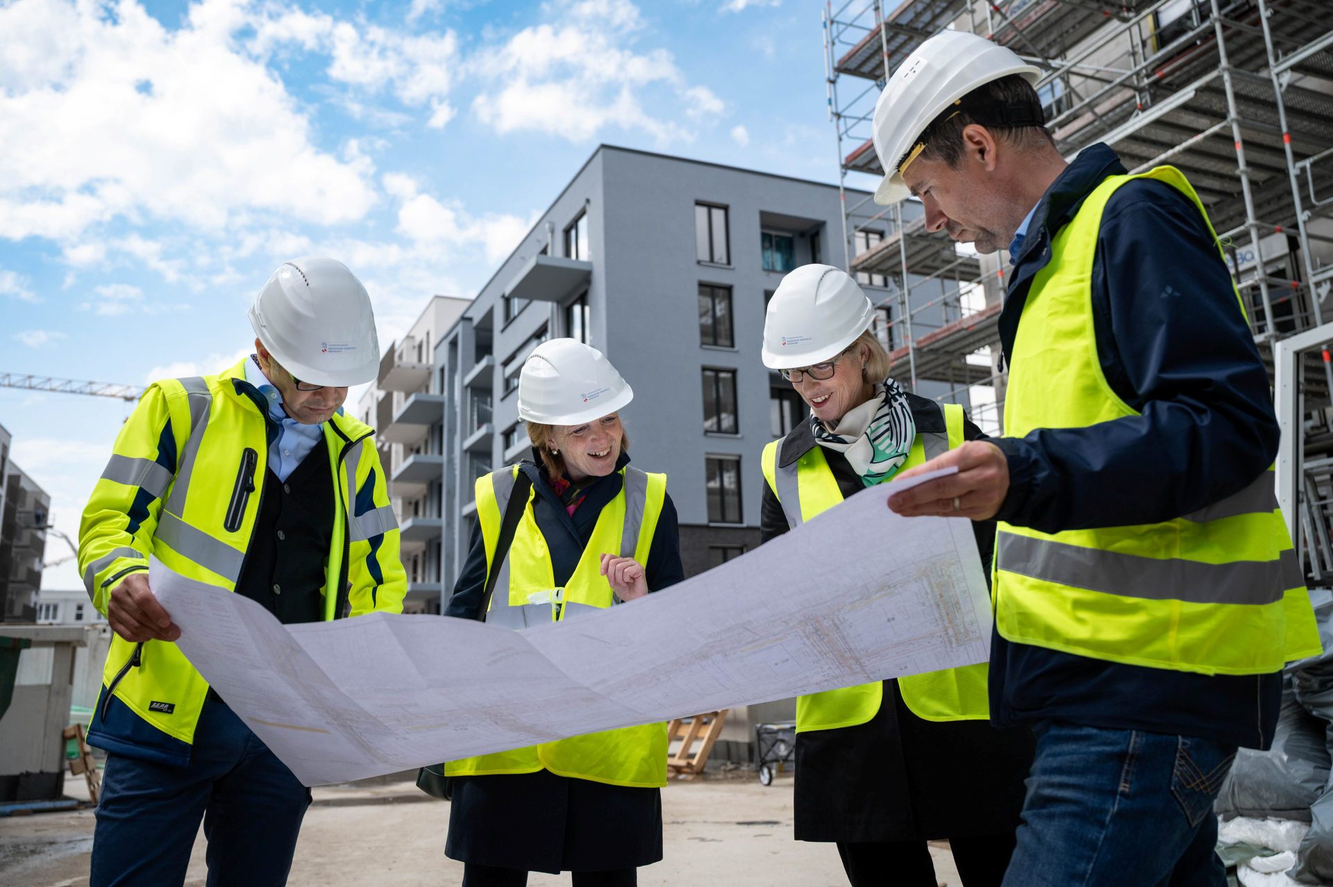 Vier Personen mit Warnweste und weißen Helmen beugen sich über einen großen Bauplan. Im Hintergrund ist Neubau Gebiet zu sehen.