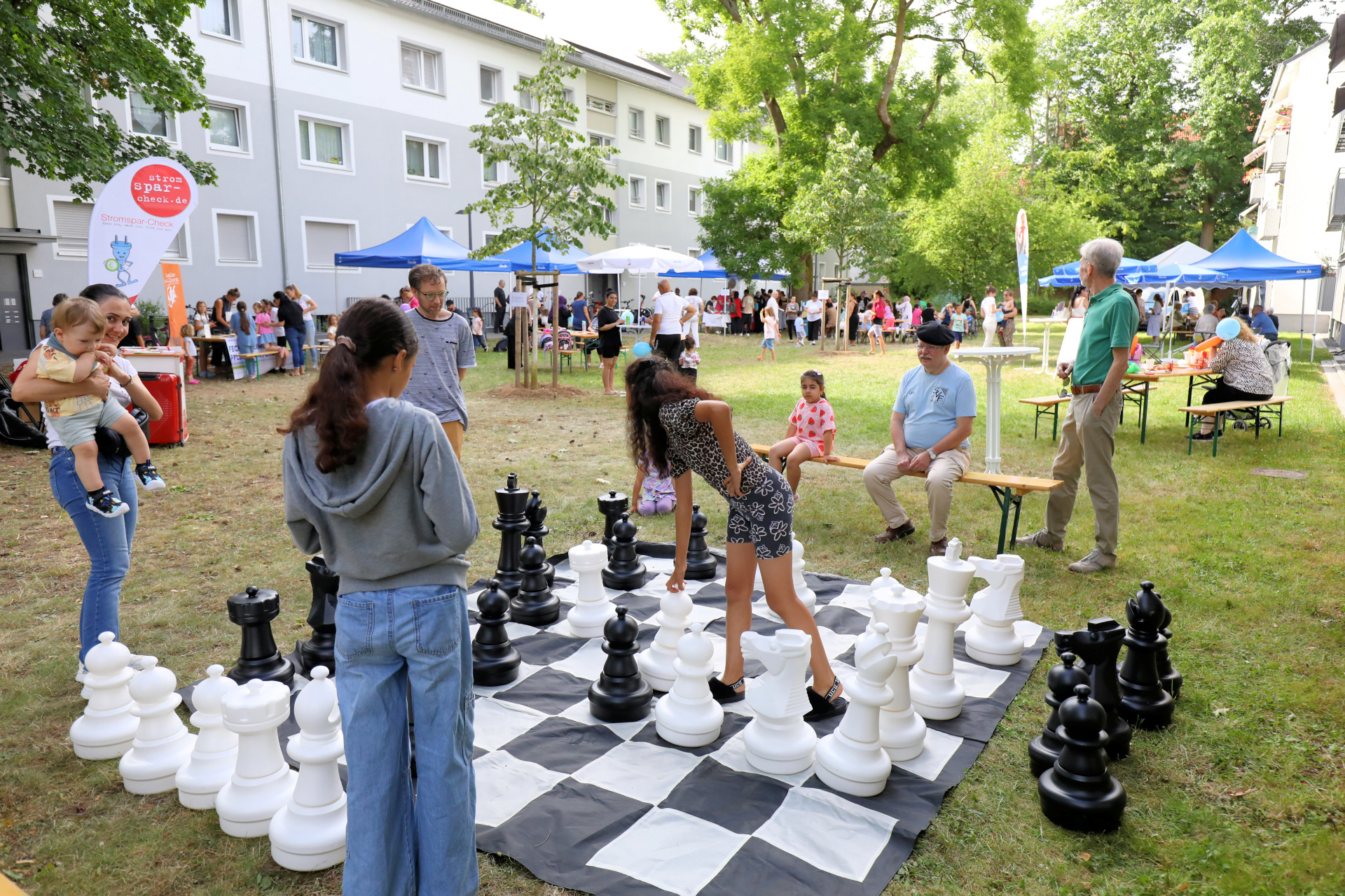 Kinder stehen um ein großes Schachbrett herum und spielen Schach. Im Hintergrund sind weitere Veranstaltungszelte zu sehen.