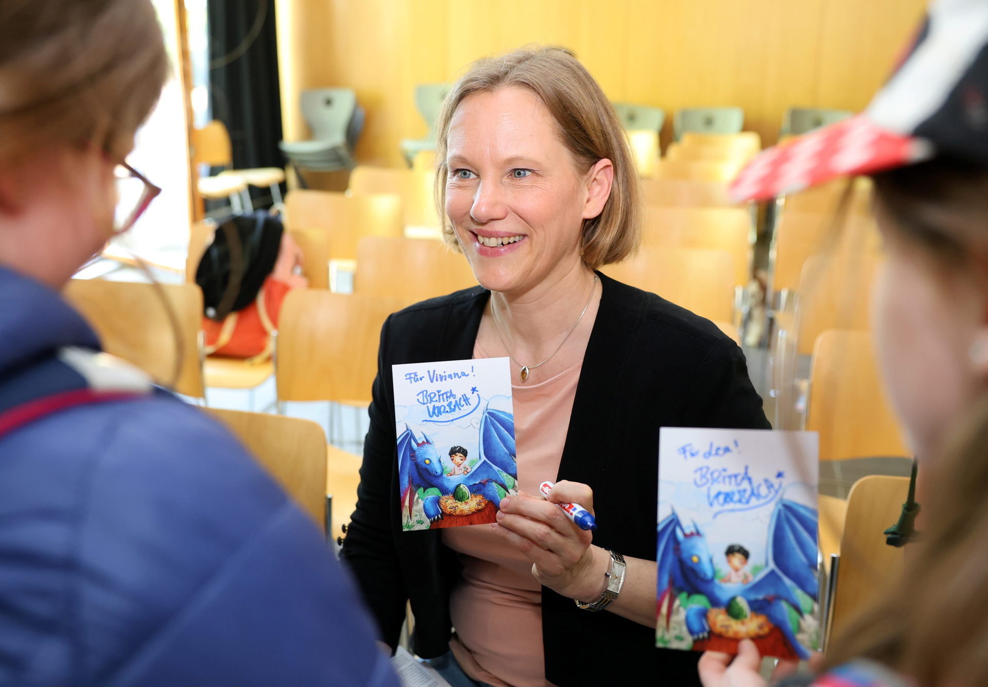 Buchautorin hält ihr Buch in der Hand und schaut seitlich in die Kamera. Im Vordergrund verschwommen zwei weitere Personen, die mit der Autorin reden
