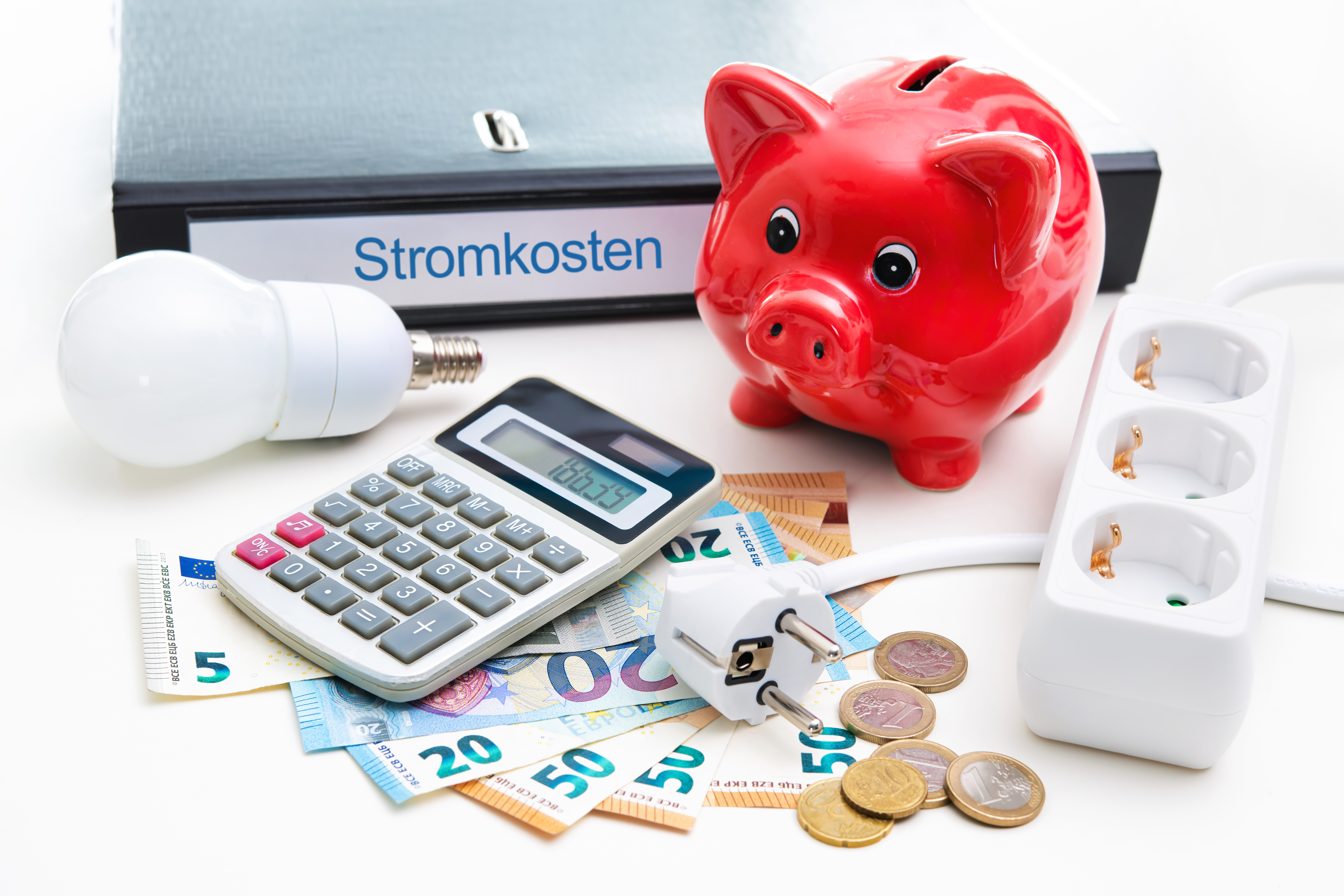Auf einem Schreibtisch liegt Bargeld, ein Taschenrechner, ein Ordner auf dem Stromkosten steht und ein rotes Sparschwein