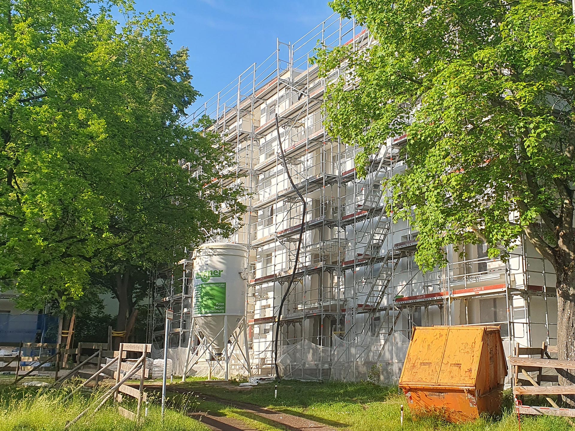 Eine Häuserzelle, die ebenfalls in ein Gerüst gehüllt ist schaut zwischen zwei großen Bäumen hervor