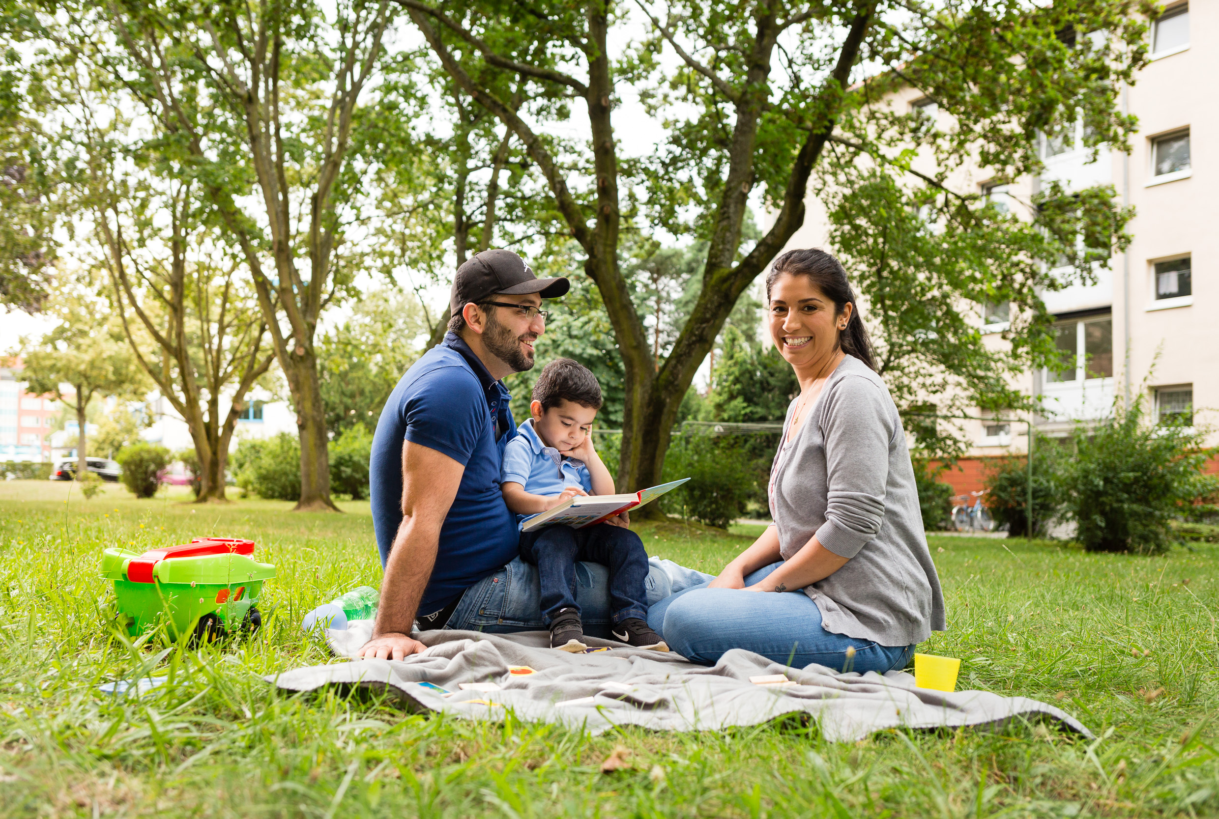 Vater, Mutter und Kind sitzen auf einer Decke auf der Wiese. Im Hintergrund sieht man Bäume und ein Haus.
